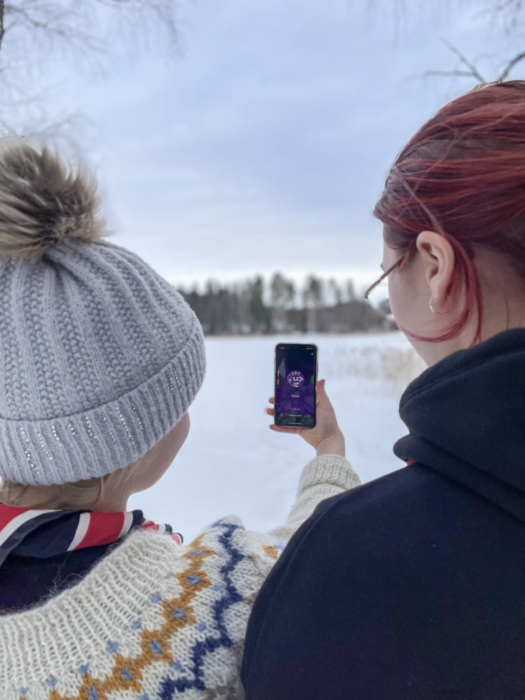 Kaksi partiolaista katsovat älypuhelimen näyttöä, jossa on auki Kompassi-sovelluksen tarpoja-otsikko. Taustalla on talvinen maisema.